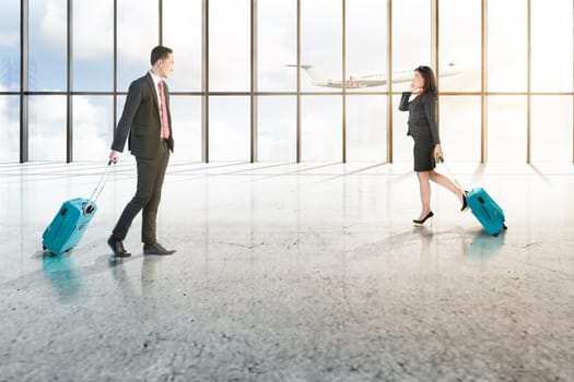 Attractive two asian business people with mobile phone and blue suitcases walking on the airport hall with window of glass and flying airplane over blue sky background