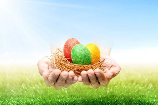 Human hands holding nest with colorful easter eggs on the grass field with sunlight and blue sky background. Happy Easter