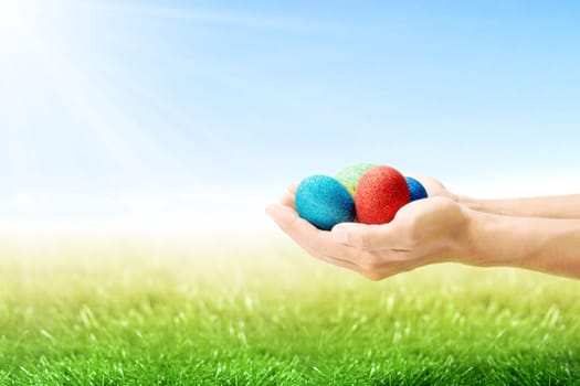 Human hands holding colorful easter eggs on the grass field with sunlight and blue sky background. Happy Easter