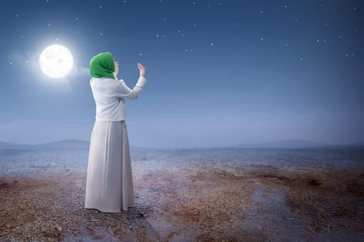 Rear view of asian muslim woman in veil raised hands and praying on the sand dune with moon and night scene background