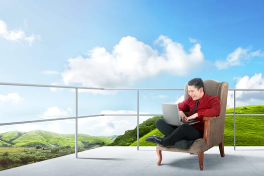 Happy asian businessman sitting on the chair and using laptop on the terrace with green landscapes view and blue sky background
