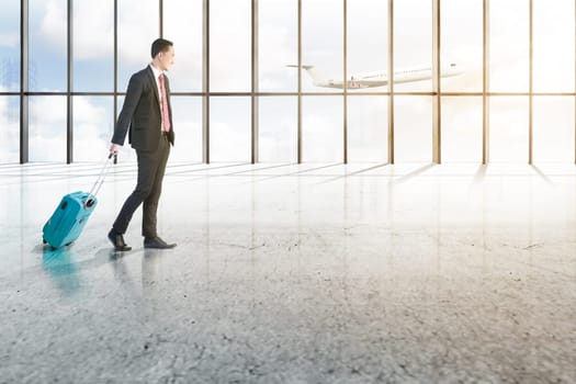 Smiling asian businessman with blue suitcase walking on the airport hall with window of glass and flying airplane over blue sky background