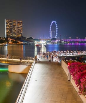 Tourists walking by Singapore embankment, illuminated Flyer, city skyline with modern architecture in background