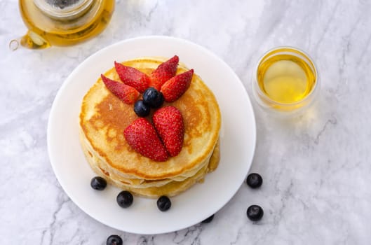 Tasty pancakes with fresh strawberry and blueberry on white plate with teapot on marble background. Table top view