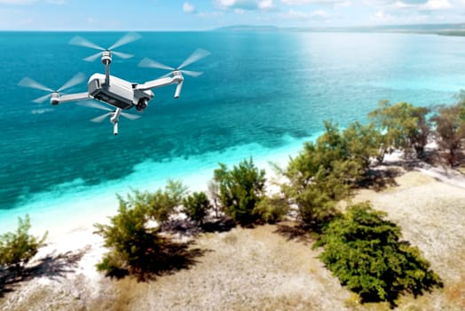 White drone with camera flying over the beach with green trees and blue ocean with blue sky background