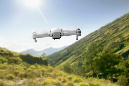 White drone with camera flying above the mountain with green grass and trees with sunlight and blue sky background