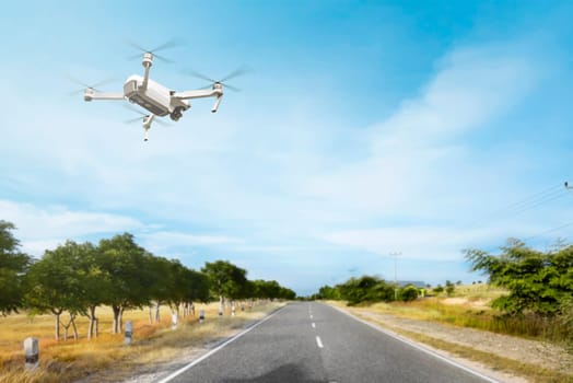 White drone with camera flying above the street with trees and grass over blue sky background