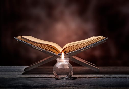 Quran open in wooden placemat and candle on wooden table over blurry background