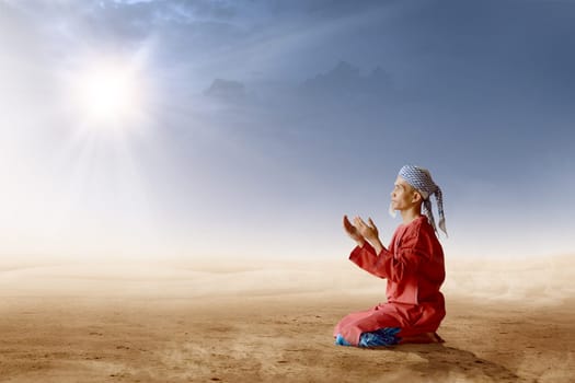 Asian muslim man with turban in his head kneeling and praying with raised hands on desert with sun rays and dark sky background