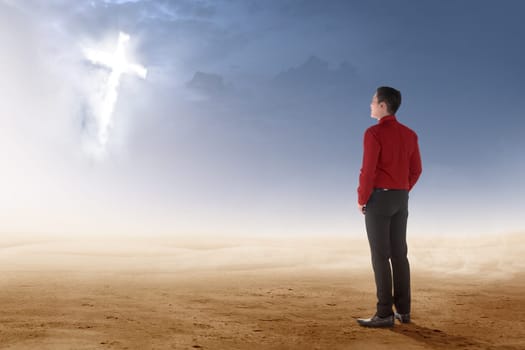 Rear view of asian businessman standing on desert and looking at glowing christian cross on the sky