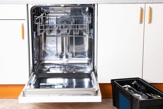 People in technician jobs. A broken built-in dishwasher with open door in a white kitchen and the service technicians toolbox.