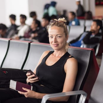 Casual tanned blond female traveler holding cell phone, passport and boarding pass while waiting to board a plane at the departure gates at the asian airport terminal.