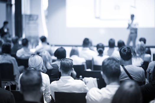 Speaker giving a talk in conference hall at business event. Focus on unrecognizable people in audience. Business and Entrepreneurship concept. Blue toned greyscale image.