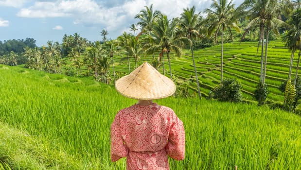 Relaxed fashionable caucasian woman wearing red asian style kimono and traditional asian paddy hat looking at beautiful green rice fields and terraces on Bali island.