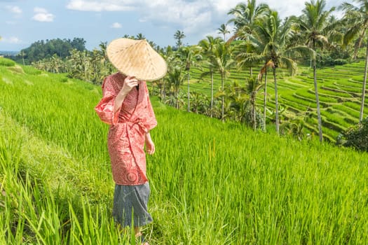 Relaxed fashionable caucasian woman wearing red asian style kimono and traditional asian paddy hat walking amoung beautiful green rice fields and terraces on Bali island.
