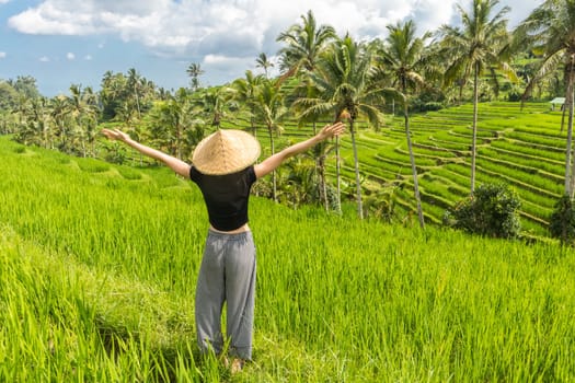 Relaxed fashionable female traveler wearing small backpack and traditional asian paddy hat, arms rised to sky, enjoying pure nature at beautiful green rice fields and terraces on Bali island.