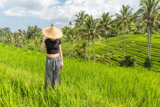 Relaxed fashionable caucasian female tourist wearing small backpack and traditional asian paddy hat looking at beautiful green rice fields and terraces on Bali island.