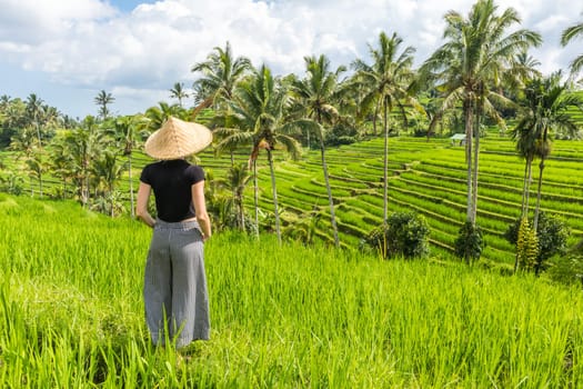 Relaxed fashionable caucasian female tourist wearing small backpack and traditional asian paddy hat looking at beautiful green rice fields and terraces on Bali island.
