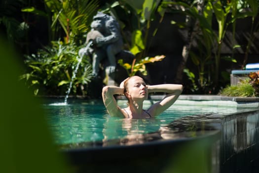 Sensual young woman relaxing in outdoor spa infinity swimming pool surrounded with lush tropical greenery of Ubud, Bali. Wellness, natural beauty and body care concept.