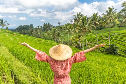 Relaxed fashionable caucasian woman wearing red asian style kimono and traditional asian paddy hat, arms rised to sky, enjoying pure nature at beautiful green rice fields on Bali island.