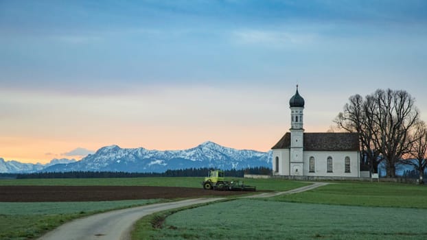 Morning farming in green Alpine field with tractor and small church on Alps snowy mountains background