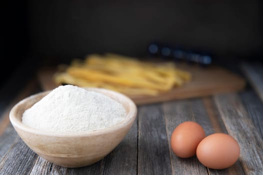 Bowl of whole wheat flour and two eggs on a rustic wooden table with pappardelle pasta in the background.