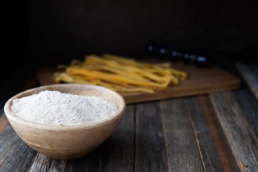 Bowl of whole wheat flour on a rustic wooden table with pappardelle pasta in the background.