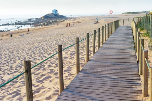 Wooden walkway, people at sandy beach in evening sunlight, Senhor da Pedra church in background by seashore, Porto, Portugal