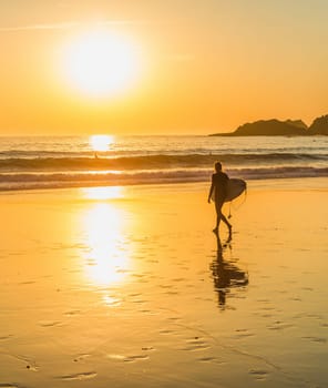 Silhouette of surfer walking to the ocean by sandy beach, scenic golden sunset over seascape, Sagres, Algarve, Portugal