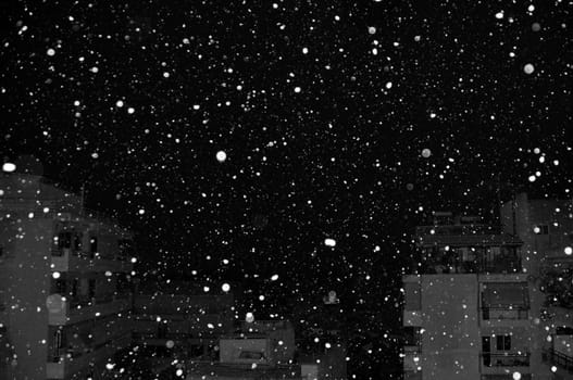 Snow falling over city buildings at night. Black and white.