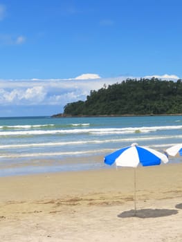 View of sunny beach with umbrella, waves and island in the background.