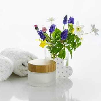 Towels next to a cream jar in front of white background and flowers in a vase.