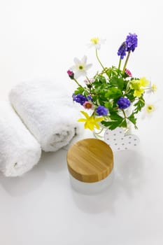 Towels next to a cream jar in front of white background and flowers in a vase.