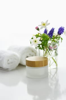Towels next to a cream jar in front of white background and flowers in a vase.