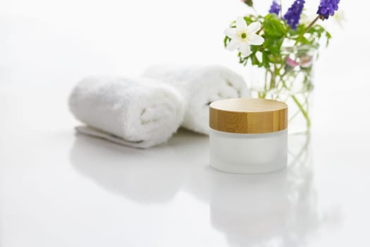 Towels next to a cream jar in front of white background and flowers in a vase.
