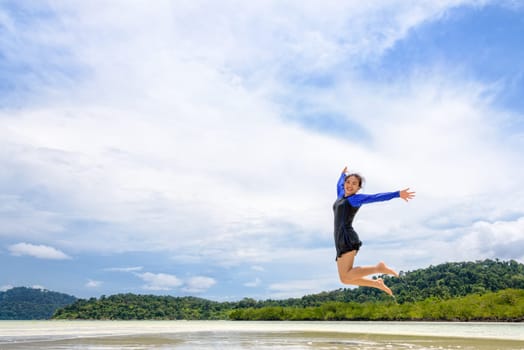 Happy asian teen girl in a swimsuit jumping fun on the beach of Ko Ra Wi on blue sky background during the sea tour vacation in summer holiday at Ko Lipe island, Tarutao National Park, Satun, Thailand