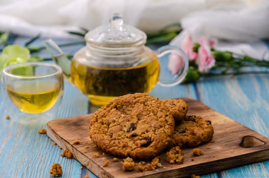 Homemade wholegrain cookies with raisin on wooden cutting board with tea and teapot on blue wooden table