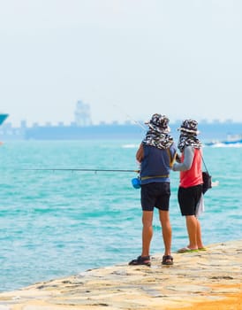 Fishermen fishing from the embankment of Singapore harbor. Commercial cargo ships in the background