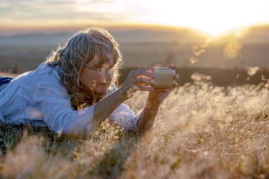 Woman lying in dry grass and taking (selfie) pictures with the smartphone in nature at sunset