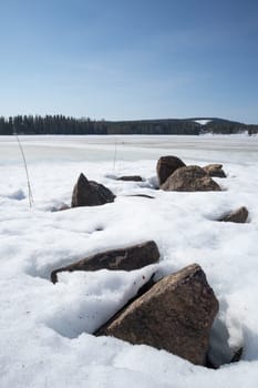 Rocks in Snow by Lake with trees in the background.