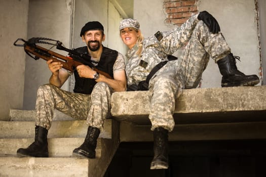 The smiling male and female army soldiers are posing in military uniform with crossbow weapon indoors as a couple or colleagues.