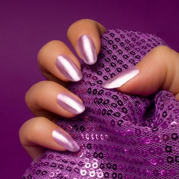 Female hand with shiny bright purple nails is holding glittered purple textile on purple background, nail care and manicure concept.