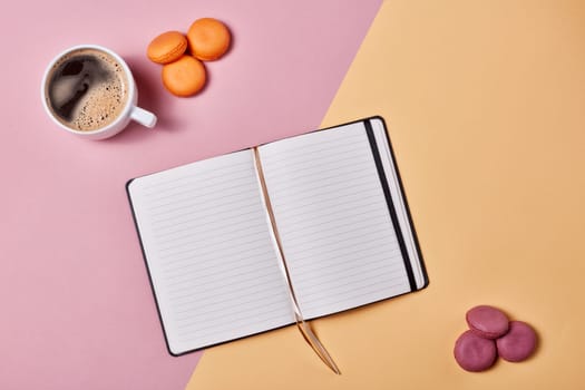 Still life with cup of coffee, delicious macaroons and notebook with blank sheet on background consisting of two colores pink and yellow. Top view with copy space. Design element for menu of cafe