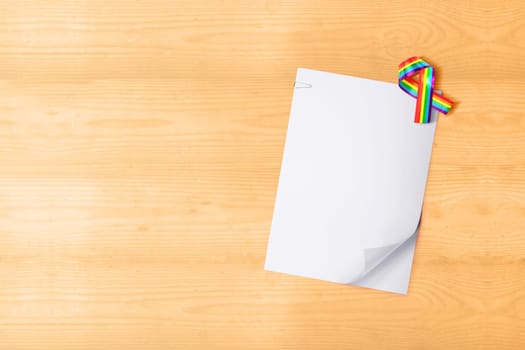 White paper with clip and ribbon with rainbow flag on wooden table. White paper for copyspace. LGBT concept
