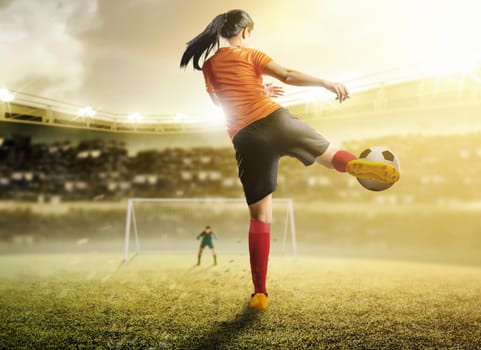 Rear view of asian football player woman in orange jersey kicking the ball in the penalty box on football field at stadium