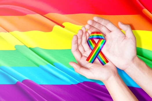 Human hands holding ribbon with rainbow flag background. LGBT concept