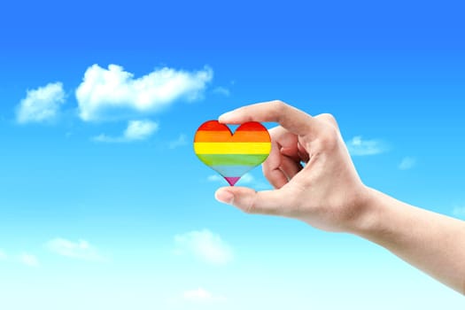 Human hands showing heart with rainbow flag over blue sky background. LGBT concept