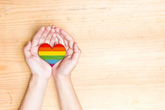 Human hands holding heart with rainbow flag over wooden table background. LGBT concept