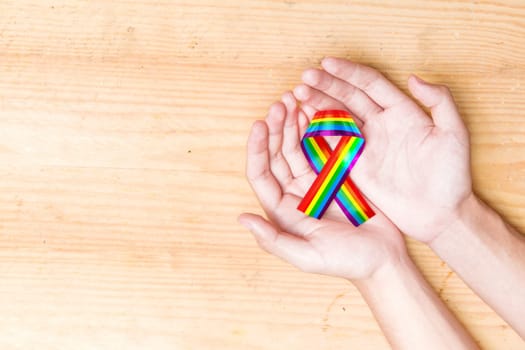 Human hands holding ribbon with rainbow flag over wooden table background. LGBT concept