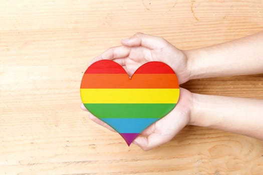 Human hands holding heart with rainbow flag over wooden table background. LGBT concept